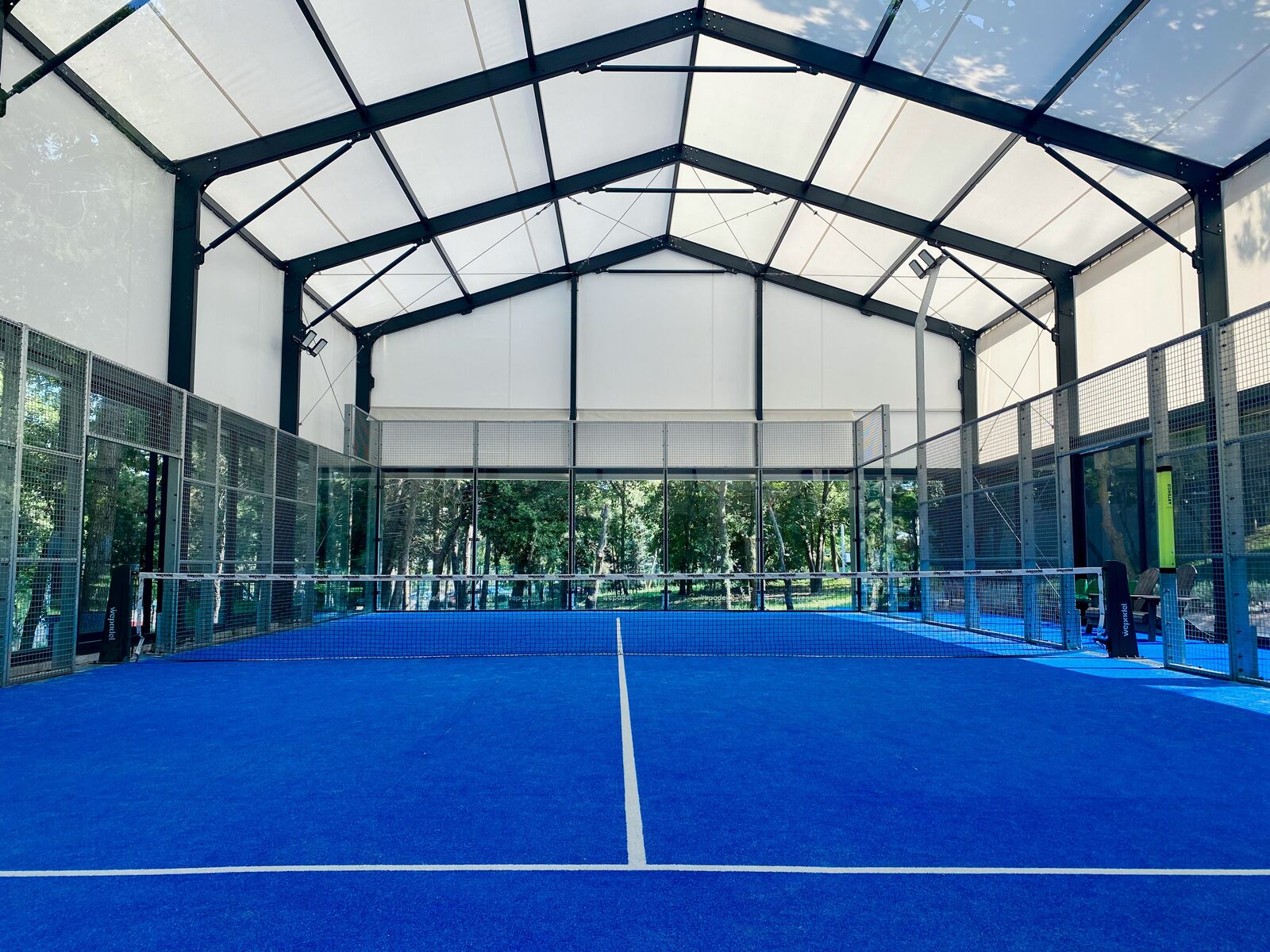 Padel courts with a roof and blue surface, surrounded by trees